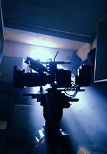 Mystical Silhouette Of A Movie Camera In The Sun's Rays Of Light From The Window. Camera On A Tripod In Standby Mode