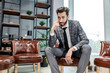 young caucasian business male in elegant stylish suit sit on leather chair in executive boardroom after business meeting with partners, personable man with beard sit near panoramic window