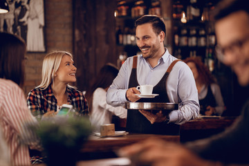 Happy waiter communicating with customers while serving them coffee in a pub.