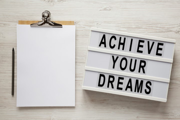 'Achieve your dreams' words on a modern board, clipboard with blank sheet of paper on a white wooden surface, top view. Overhead, from above, flat lay.