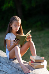 charming little girl with long brown hair reads book outdoor sitting on tree in summer park or in a 