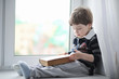 © alexkich - The little boy is reading a book. The child sits at the window and prepares for lessons. A boy with a book in his hands is sitting on the windowsill.