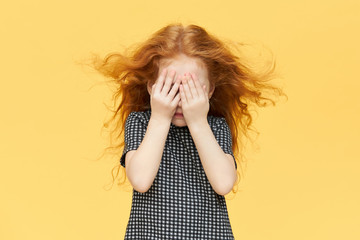 Isolated shot of timid little girl being shy, covering eyes. Red haired female child hiding tears behind her hands, crying. Caucasian kid playing seek and hide against blank yellow wall background