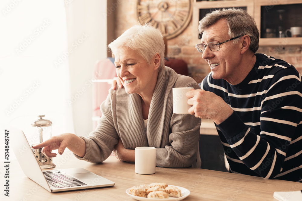 Retired couple browsing on laptop, enjoying tea at kitchen - obrazy, fototapety, plakaty 