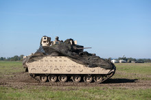 Fighting Vehicle Used By US Army Is Moving On Field And Battlefield. Machine Has Camouflage Over Turret. Unrecognizable Soldier Is On The Top. Pannign Shot With Slightly Blurred Background.