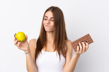 Wall Mural - Young woman over isolated white background having doubts while taking a chocolate tablet in one hand and an apple in the other