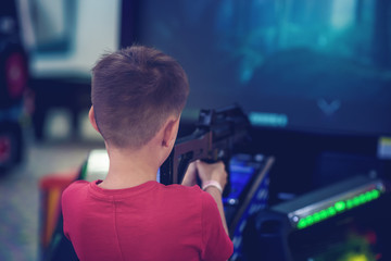 Wall Mural - Cute European boy in a red t-shirt is playing with automatic slot-gun in shooting gallery. Back view.