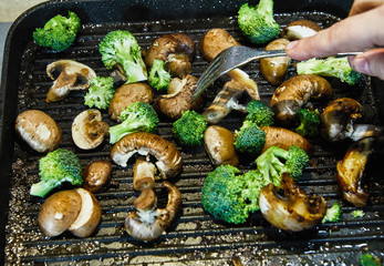 Poster - Fresh champignon mushrooms and porcini mushrooms are fried on a board along with broccoli in butter. Female hand with a fork checks readiness and mixes. Healthy eating concept