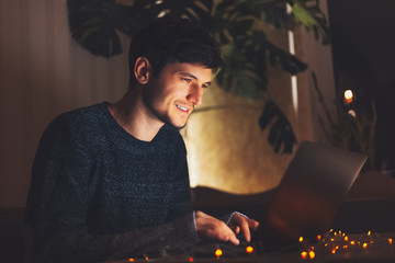 Night portrait of young cheerful guy working on laptop in dark room with garlands on table at home.