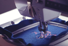 View On Machine Head And Embroidery Hoop Of A Modern Sewing Machine That Stitches A Magnolia With Light Pink Yarn On Classic Blue Boiled Wool - Moody Toned Style - Background And Foreground Blurry