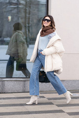 Wall Mural - European model girl in a beige oversized down jacket, knitted sweater, jeans flared with a handbag and in glasses is walking along the street near glass windows. Life style