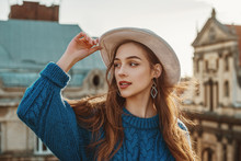 Outdoor Close Up Fashion Portrait Of Young Happy Brunette Woman With Long Natural Hair. Model Wearing Blue Sweater, Stylish White Hat, Trendy Earrings, Posing At Sunset, In European City. Copy Space