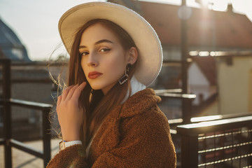 Young elegant fashionable brunette woman, model wearing stylish white hat, wrist watch, brown faux fur coat, posing at sunset, in European city. Copy empty space for text