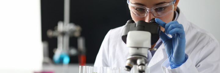 Adult caucasian beauty woman chemist in protective glasses looking at microscope against chemistry lab background. Medical analysis processing concept