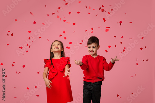 Fotografía Happy romantic little couple child girl and boy stand in the rain of flying rose petals on pink background