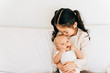 © twinsterphoto - Caring Asian girl in casual wear hugging small brother embracing newborn baby with opened mouth smelling toddler sitting on bed in house