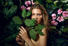 A Beautiful Blue-eyed Long-haired Brown-haired Woman Stands On A Background Of Exotic Plants And Pink Orchids.