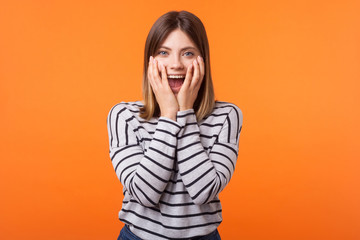 Wall Mural - Portrait of shocked young woman with brown hair in long sleeve striped shirt standing, looking with amazement and hiding face in arms, unexpected news. indoor studio shot isolated on orange background