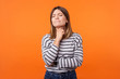 Portrait of ill woman with brown hair in long sleeve striped shirt standing touching neck and frowning from pain, suffering sore throat, flu symptom. indoor studio shot isolated on orange background