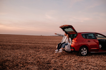 Young Happy Couple Dressed Alike in White Shirt and Jeans Sitting at Their New Car Trunk, Beautiful Sunset on the Field, Vacation and Travel Concept