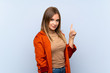 © luismolinero - Teenager girl with coat over isolated blue background showing and lifting a finger in sign of the best