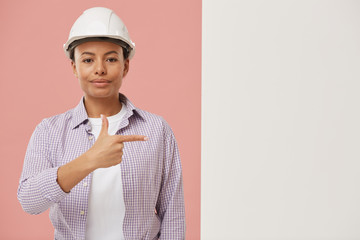 Waist up portrait of beautiful female worker looking at camera and pointing at blank white sign against pale pink background, copy space