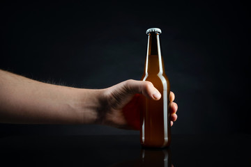 Wall Mural - Men holding cold unopened bottle of beer with cap on black background. Hand holding glass of refrigerated wheat or lager beer on dark background