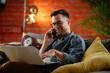 © JustLife - Young man working from home. Businessman at home on sofa with laptop and phone.