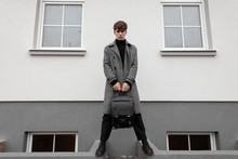 Fashion Model Of A Stylish Young Man In An Elegant Plaid Coat With A Leather Black Vintage Backpack In Trendy Boots In The City Near A White Wall. Modern Handsome Guy. Autumn-winter Style. Menswear.