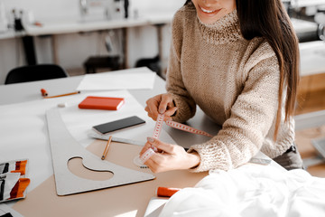 Poster - One woman with beautiful smile sitting at the table with tailoring accessories in hands. Preparing to sew a new item of clothing for a private client