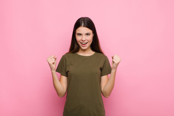 young pretty woman shouting aggressively with an angry expression or with fists clenched celebrating success against pink wall