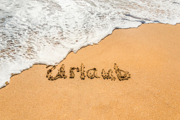 Hand written text in German Urlaub (English translation holidays) on the golden beach sand with coming wave