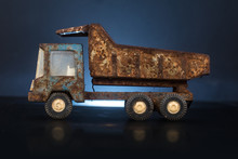 Side Of An Old, Dirty And Very Rusty Tin Metal Model Toy Of A Dump Truck In Blue And Grey Color In A Black And Blue Backgorund On Reflective Surface. One Wheel Is Missing.