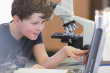 Boy Student Conducting Scientific Experiment At Microscope And Computer In Laboratory Classroom
