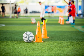 football and marker cones on green artificial turf with blurry soccer team training, blurry kid soccer player jogging between marker cones and control ball with soccer equipment in soccer academy.