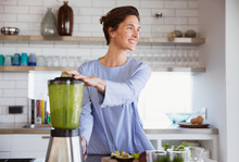 Smiling brunette woman making healthy green smoothie in blender in kitchen