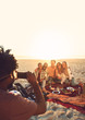 © Trevor Adeline/Caia Image - Young man camera phone photographing friends enjoying picnic on sunny summer beach