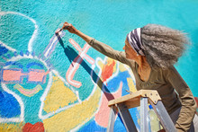 Senior Woman Painting Vibrant Mural On Sunny Wall