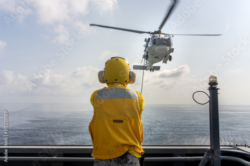Helicopter deck officer give hand signal to Sikorsky S-70 Sea Hawk helicopter hovering above helicopter deck of Navy ship to perform hot refueling while flying at sea.
