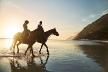 Young Women Horseback Riding In Sunny Sunset Ocean Surf