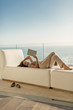 © Tom Merton/Caia Image - Woman using digital tablet relaxing laying on chaise lounge on luxury balcony with ocean view