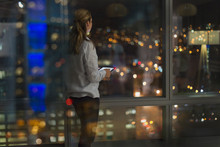 Pensive businesswoman with digital tablet working late, looking out urban office window at night