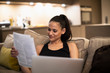 © Sam Edwards/Caia Image - Smiling woman reading paperwork and using laptop on sofa