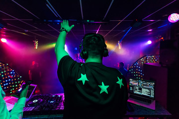 back of a male DJ with a raised hand in headphones at an electronic concert