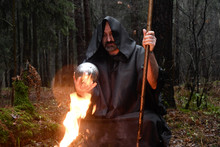 A Black-robed Monk Sits By A Fire In The Forest And Looks Into A Divination Ball. Mysticism And Magic, Sorcerers And Witches.