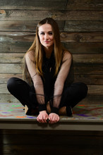 The Concept Of A Glamorous Portrait. A Full-length Photo Of A Cute Brunette With Excellent Makeup In Dark Clothes Is Sitting On A Stand On A Wooden Background In The Home Interior.