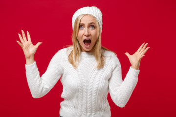 Wall Mural - Shocked young woman in white sweater, hat isolated on red background, studio portrait. Healthy fashion lifestyle, cold season concept. Mock up copy space. Screaming keeping mouth open spreading hands.