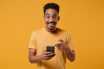 Funny young african american guy in casual t-shirt posing isolated on yellow orange background, studio portrait. People lifestyle concept. Mock up copy space. Pointing index finger on mobile phone.