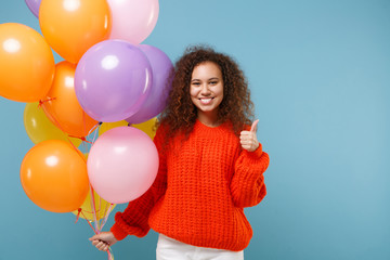 Wall Mural - Smiling african american girl in orange knitted clothes isolated on pastel blue background. Birthday holiday party, people emotions concept. Celebrating holding colorful air balloons showing thumb up.