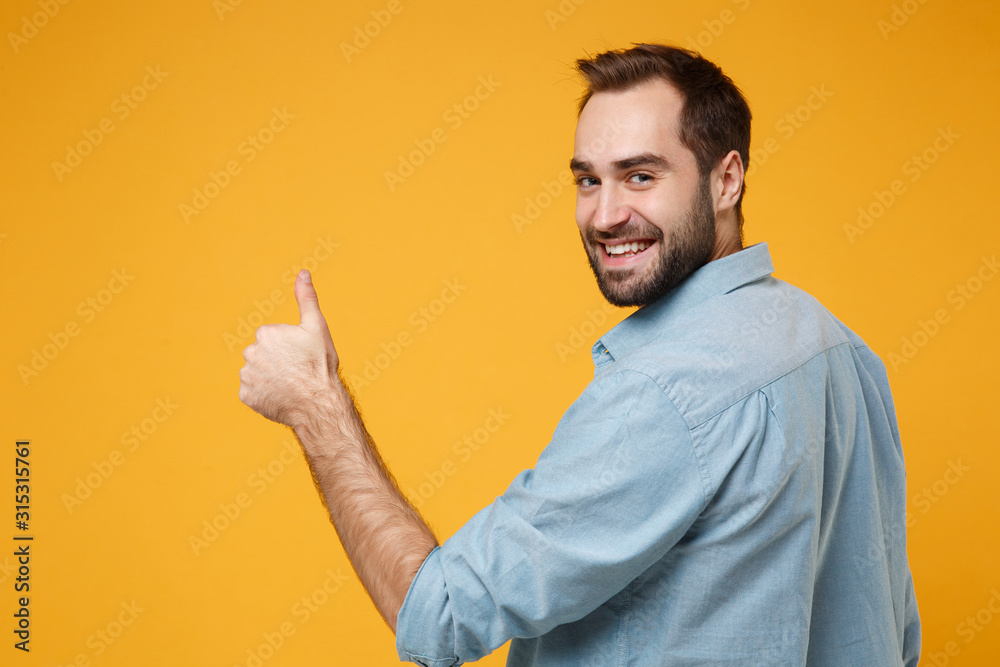 Back rear view of smiling young bearded man in casual blue shirt posing isolated on yellow orange background in studio. People lifestyle concept. Mock up copy space. Looking back, showing thumb up. - obrazy, fototapety, plakaty 